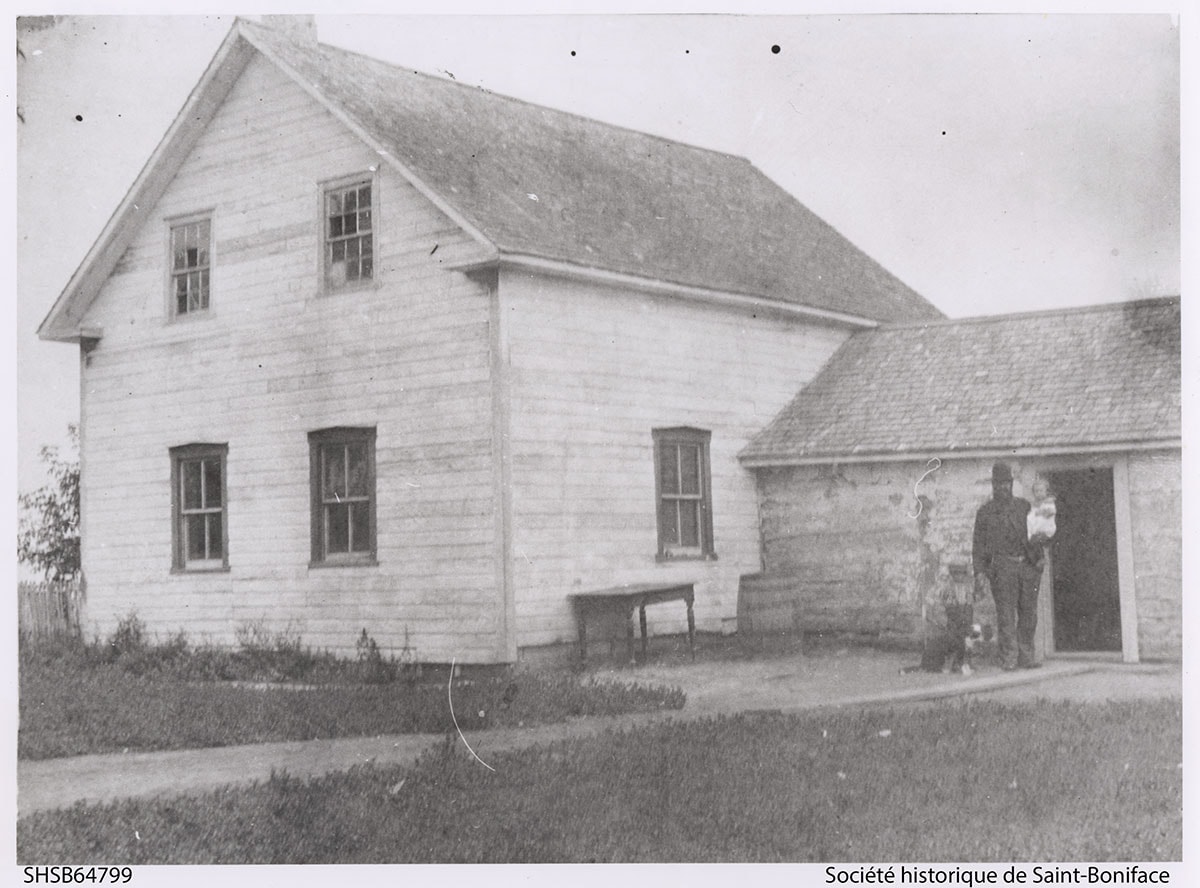 Photographie de la Maison Riel prise aux environs de 1891-1892. On y voit debout devant Joseph Riel et dans ses bras un enfant ainsi qu'un chien à ses côtés. (David est né en 1888 et Sara en 1890). SHSB 64799